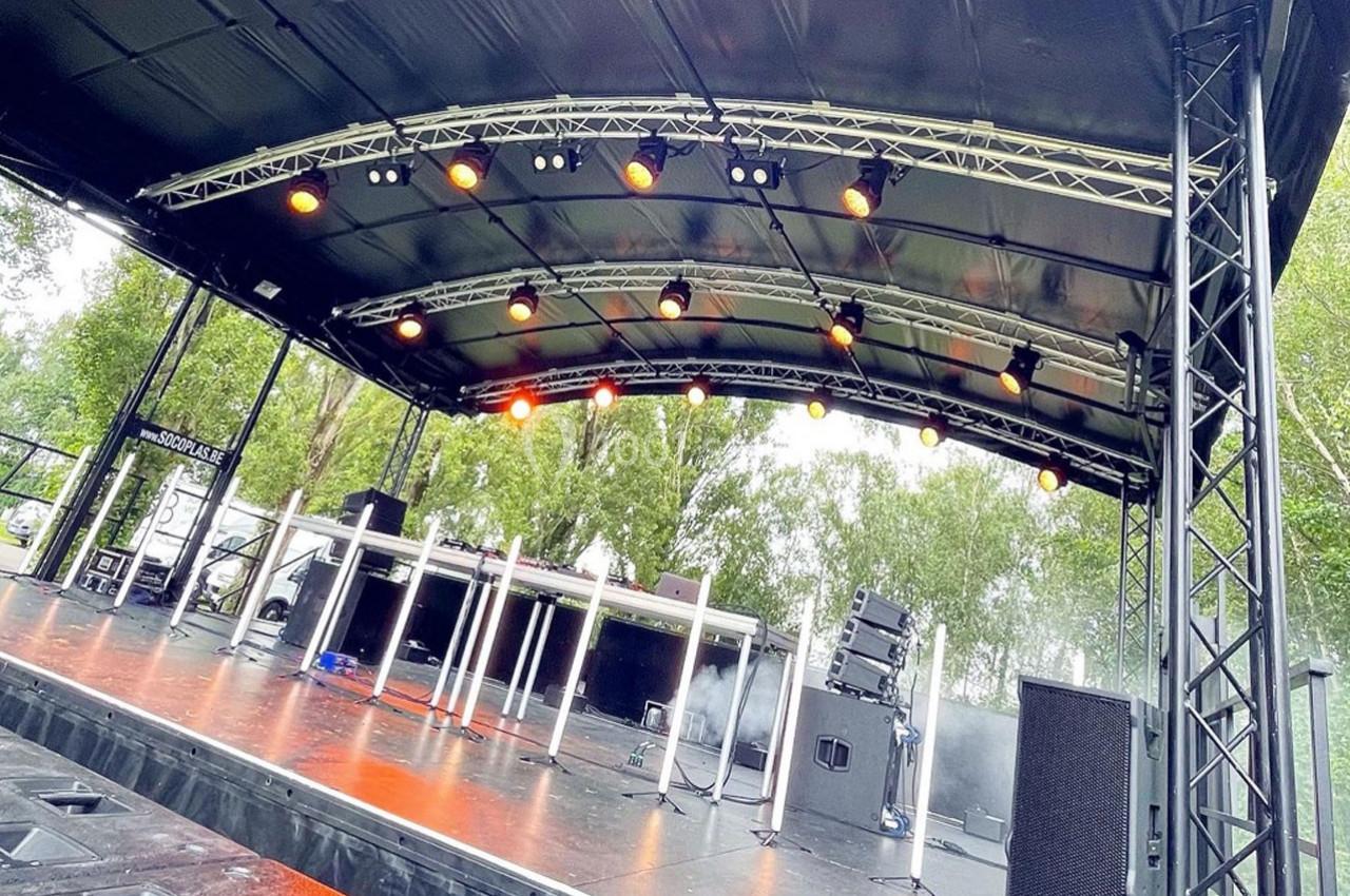 Scène extérieure avec structure métallique, éclairages suspendus et équipements audio, entourée d'arbres.