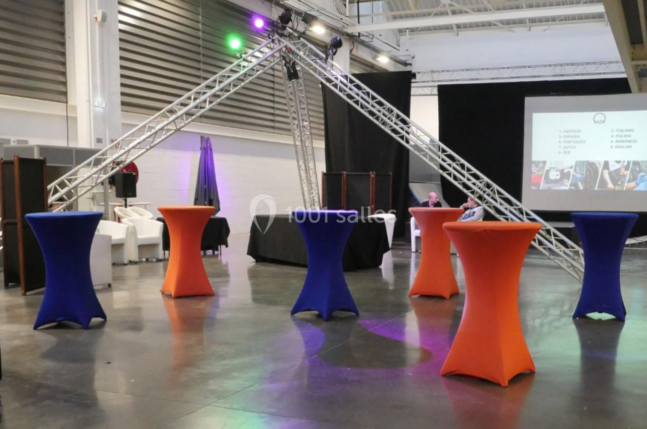 Salle d'événement avec tables hautes colorées, structure métallique éclairée et écran de projection en arrière-plan.