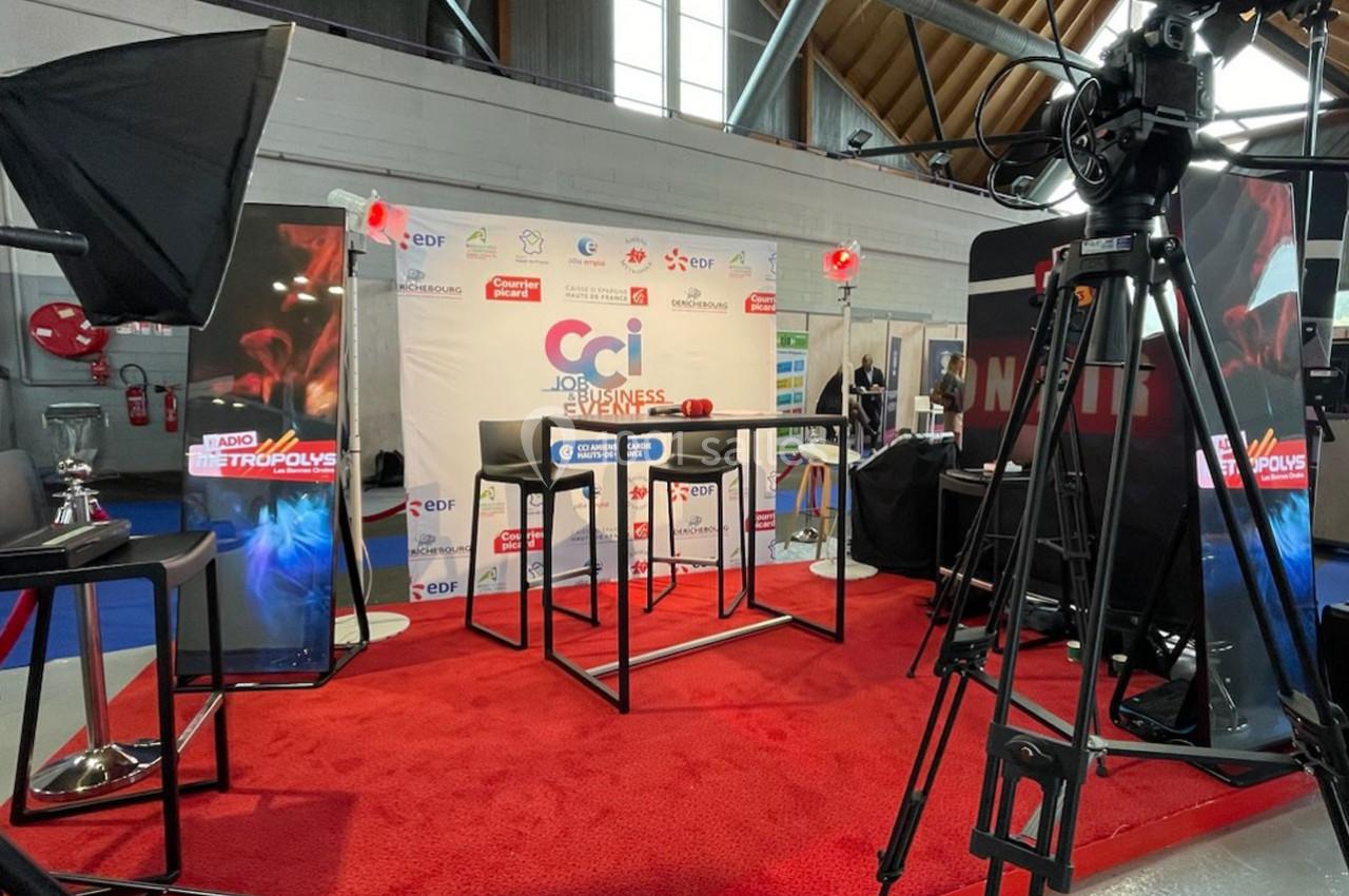 Stand d'exposition avec table haute, éclairages, fond promotionnel et équipements audiovisuels sur tapis rouge.