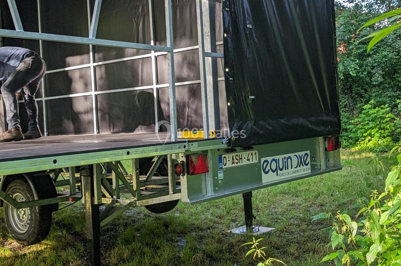 Remorque ouverte avec structure métallique et bâche noire, stabilisée sur un terrain herbeux près de végétation.