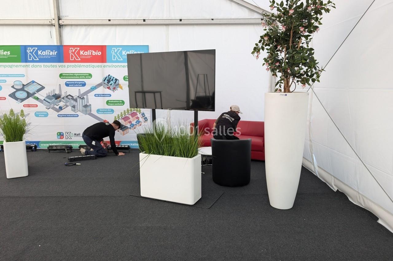 Un stand sous tente avec un écran, des plantes en pots, un canapé rouge et deux personnes installant du matériel.