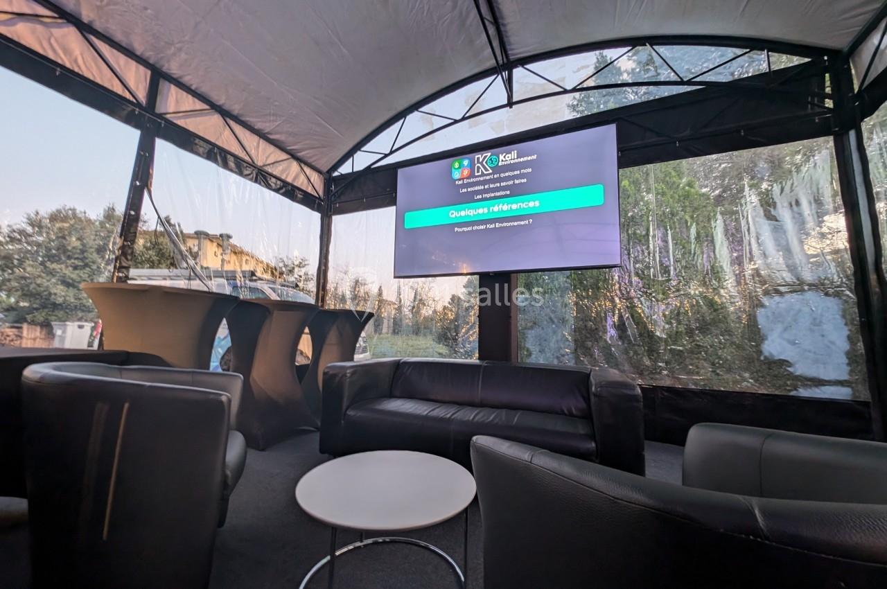 Espace lounge sous une tente avec canapés, table basse et écran affichant une présentation.