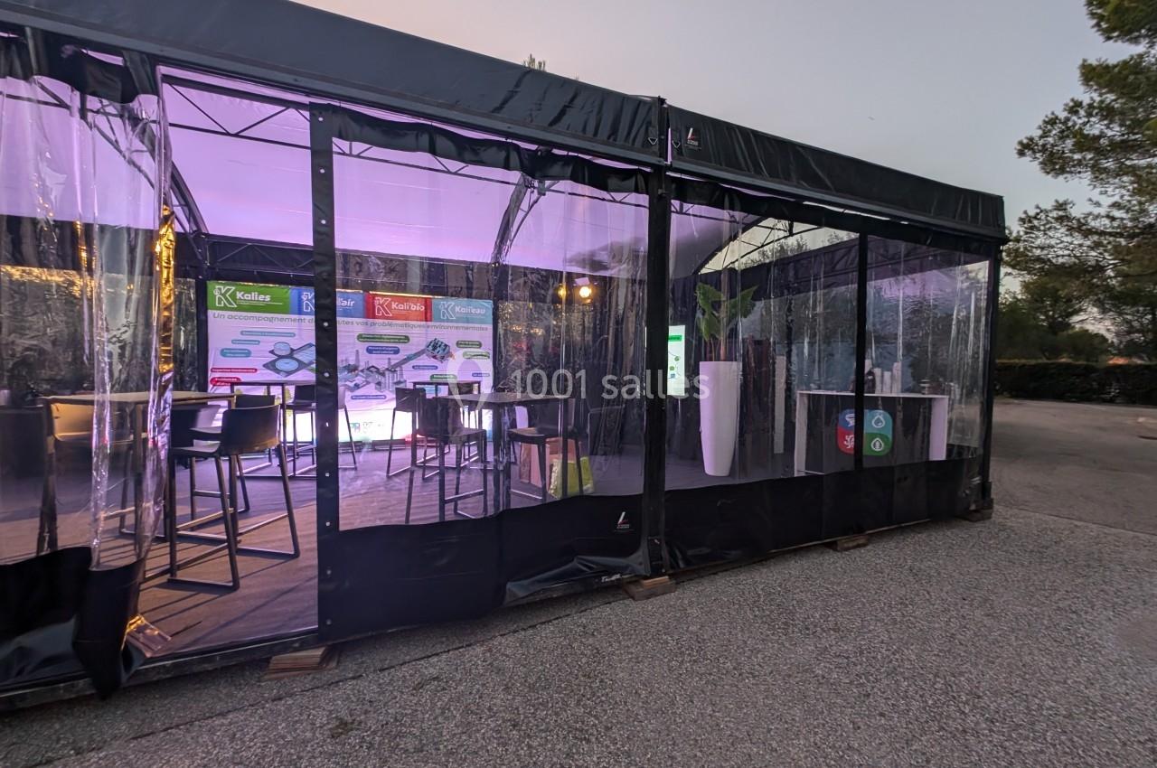 Vue extérieure d'un stand sous tente avec parois transparentes, éclairage violet et panneaux d'information visibles à l…