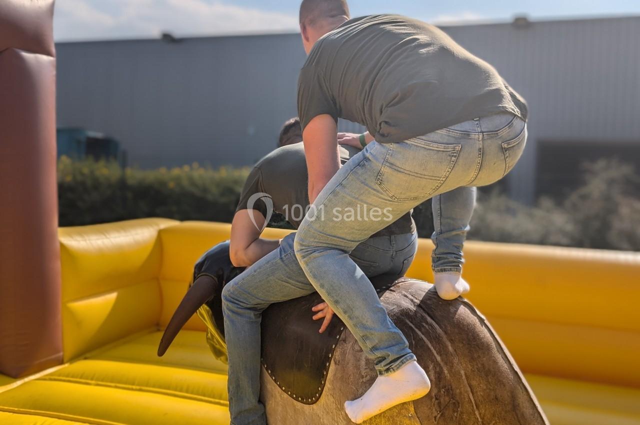 Deux personnes en jeans tentent de monter sur un taureau mécanique dans une structure gonflable en extérieur.