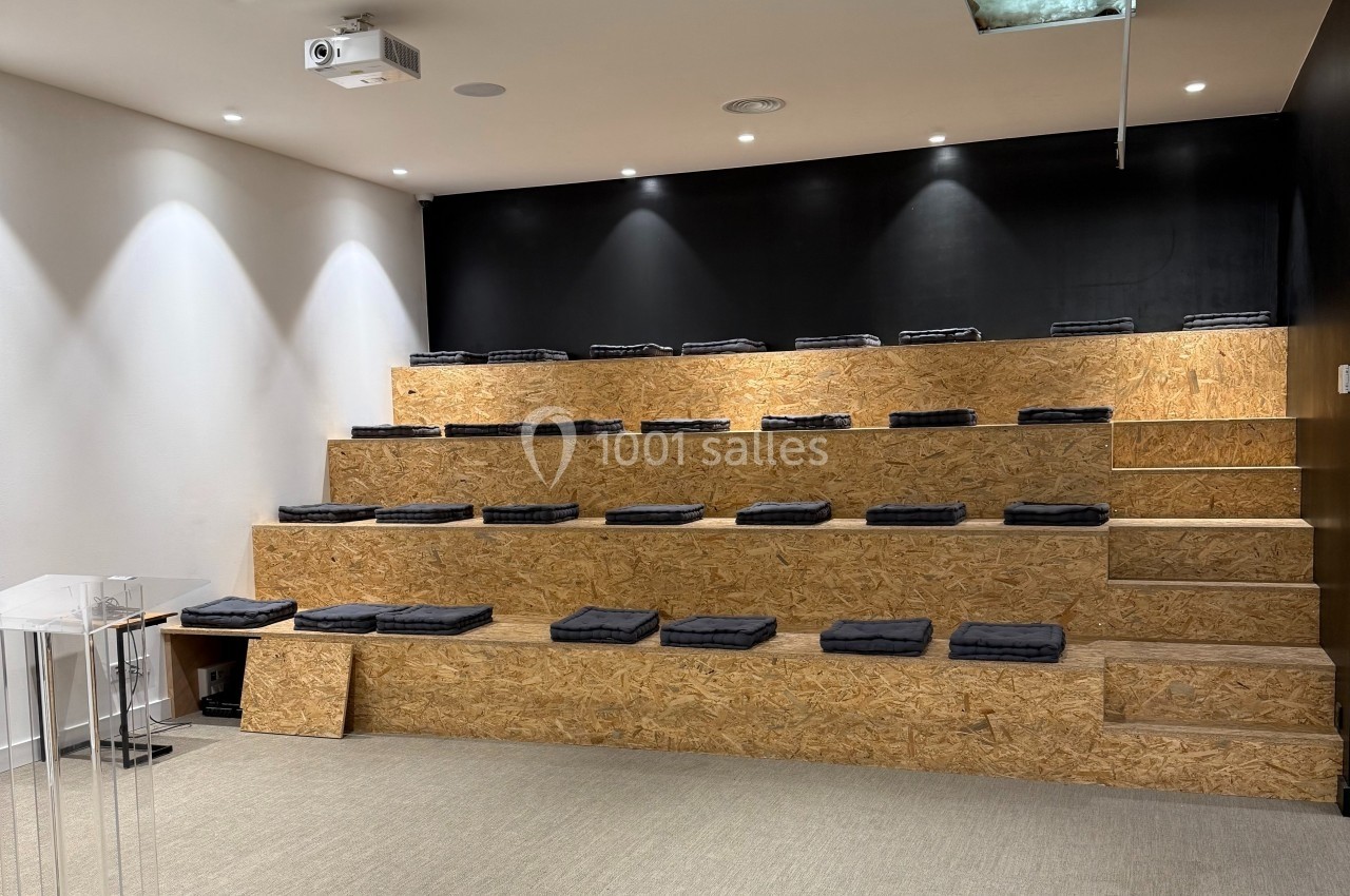 Salle avec gradins en bois, coussins noirs disposés sur les sièges, mur noir et plafond équipé d'un projecteur.