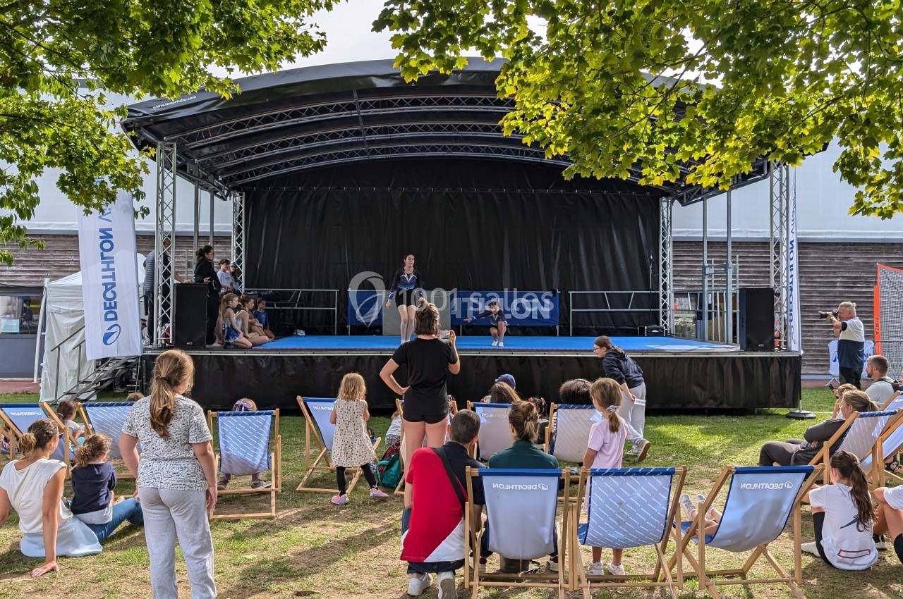 Des spectateurs assis devant une scène en plein air où se déroule une présentation, entourée d'arbres et d'installations.