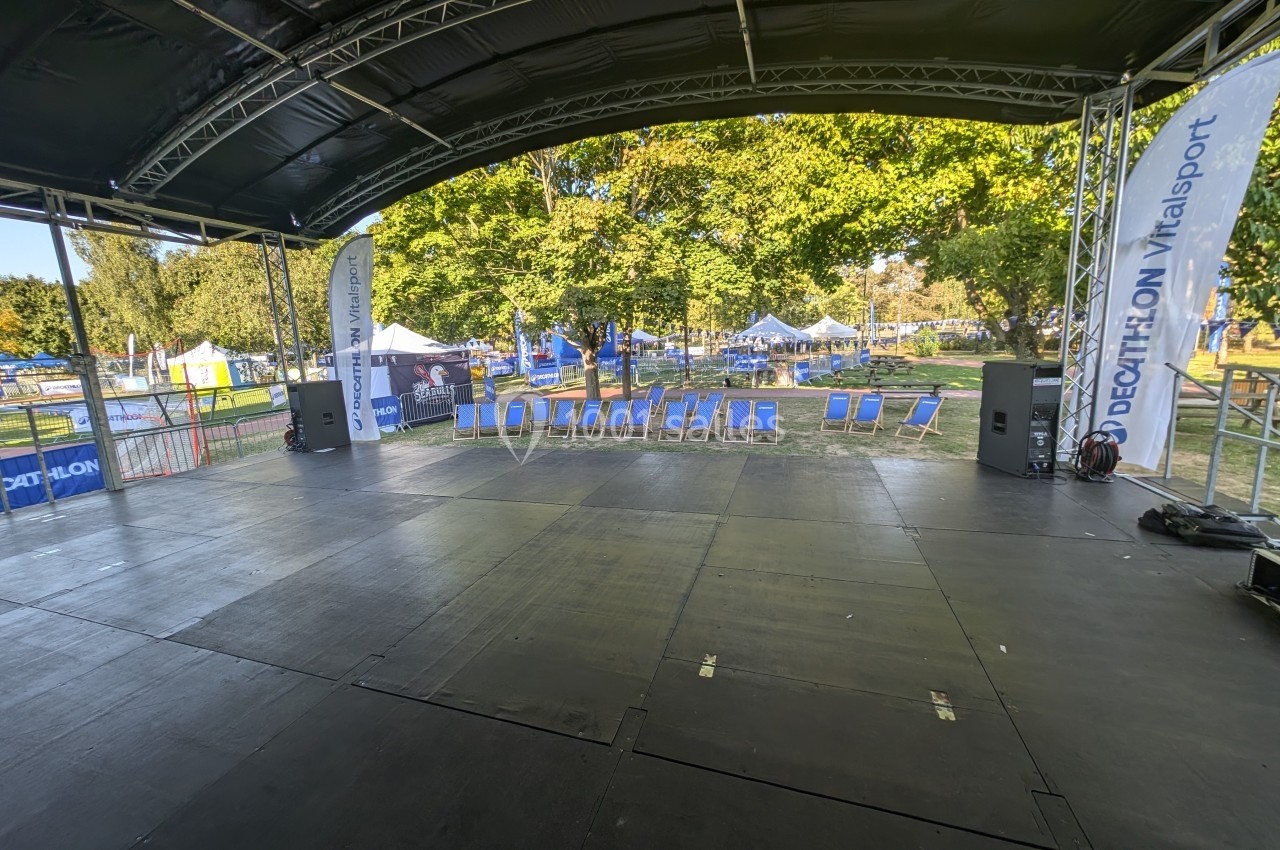 Scène vide sous un chapiteau, entourée de chaises longues et de tentes dans un parc arboré.