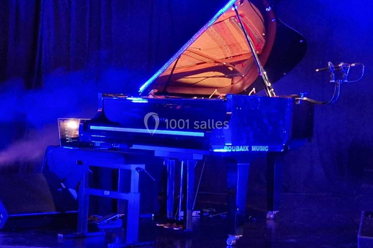 Piano à queue noir éclairé par une lumière bleue, avec un micro installé à proximité, sur une scène sombre.
