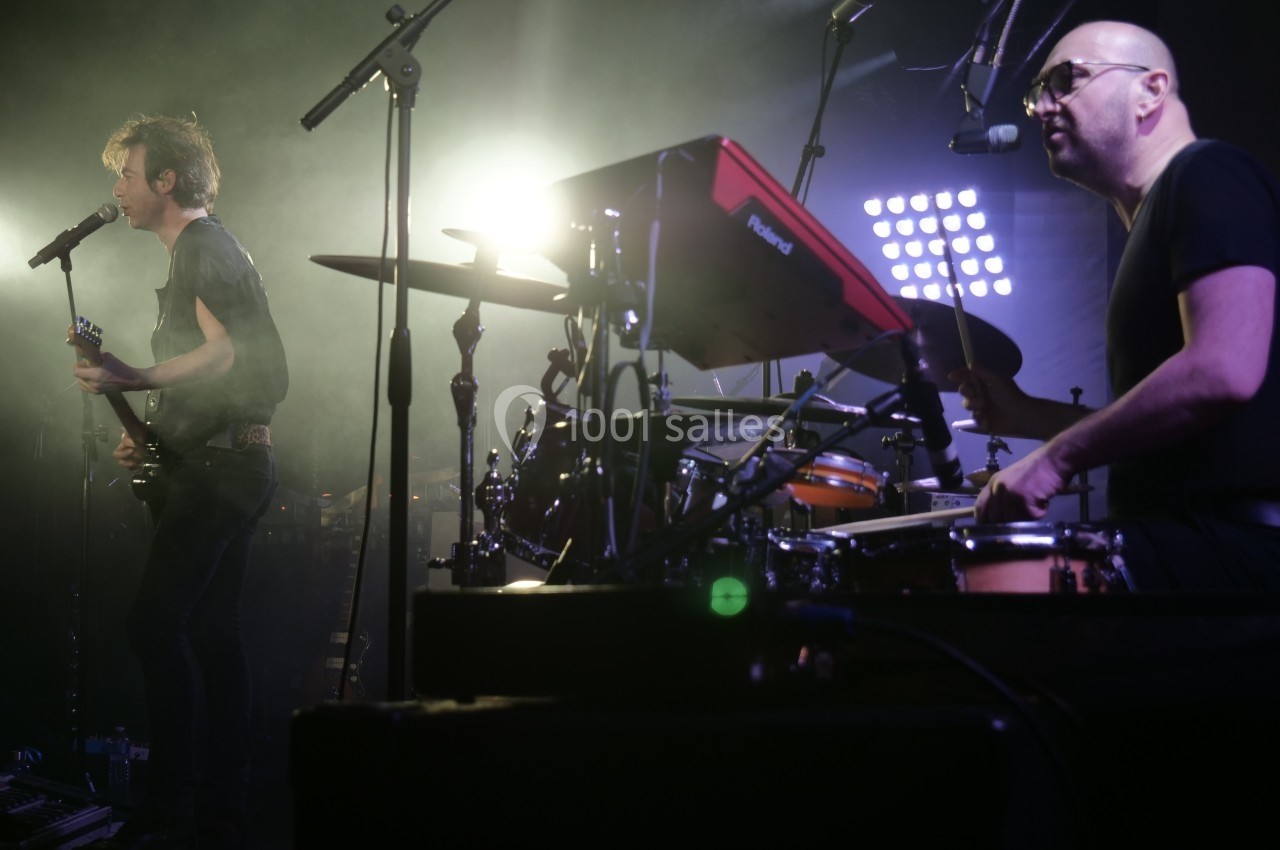 Un chanteur joue de la guitare et chante sur scène tandis qu'un batteur joue à ses côtés sous des lumières de concert.