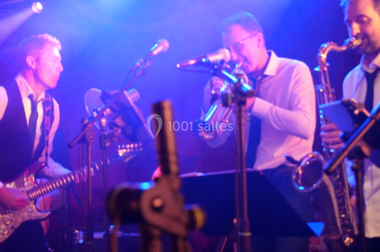 Musiciens jouant de la guitare, de la trompette et du saxophone sur une scène éclairée par des lumières colorées.