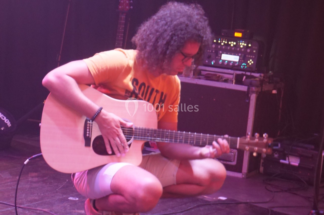 Un musicien accroupi joue de la guitare acoustique sur une scène avec des équipements en arrière-plan.