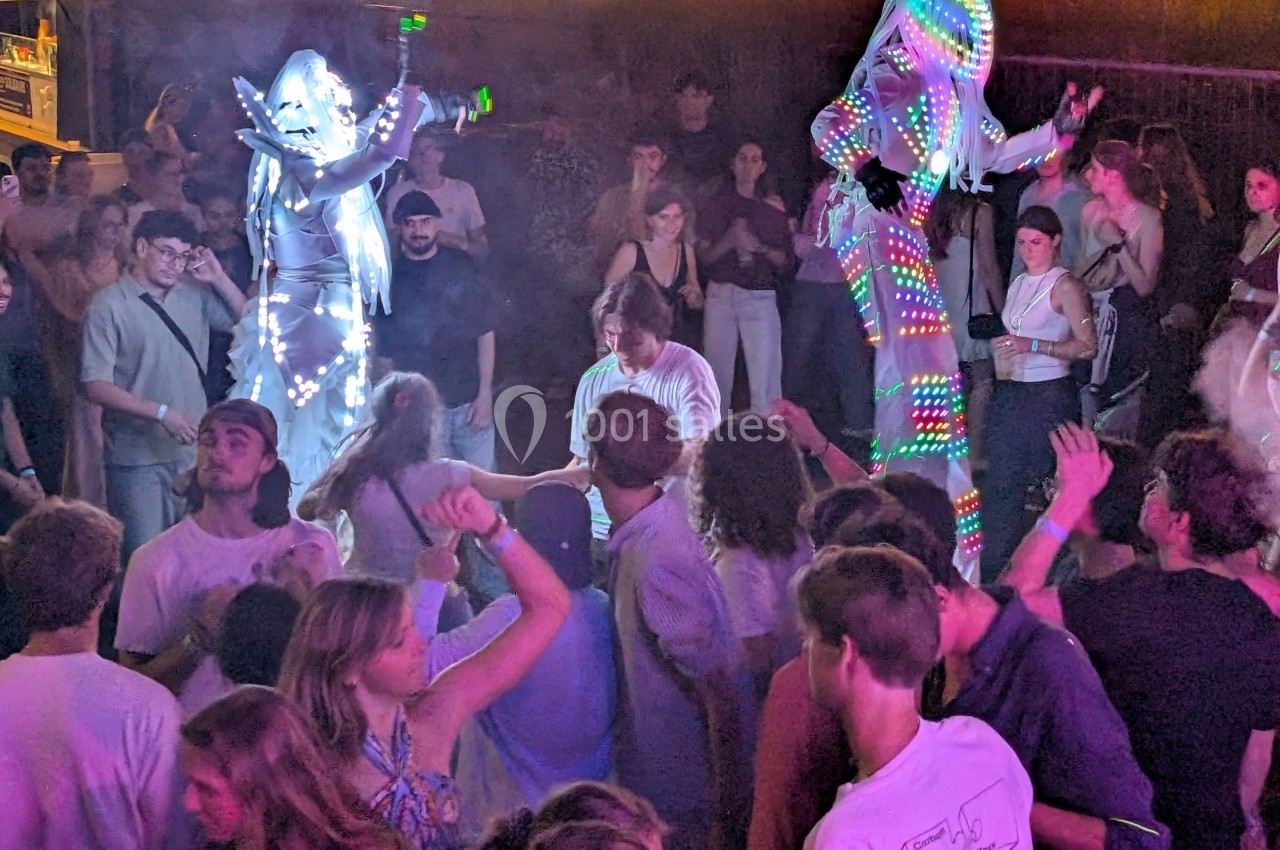 Des artistes en costumes lumineux dansent sur des échasses au milieu d'une foule lors d'une soirée festive.