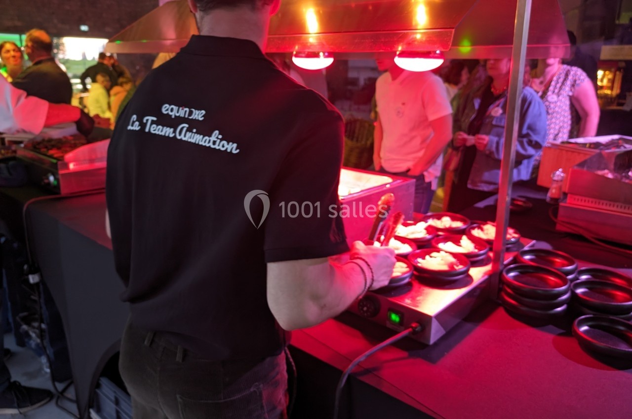 Un cuisinier prépare des plats sous une lampe chauffante lors d'un événement avec des personnes en arrière-plan.