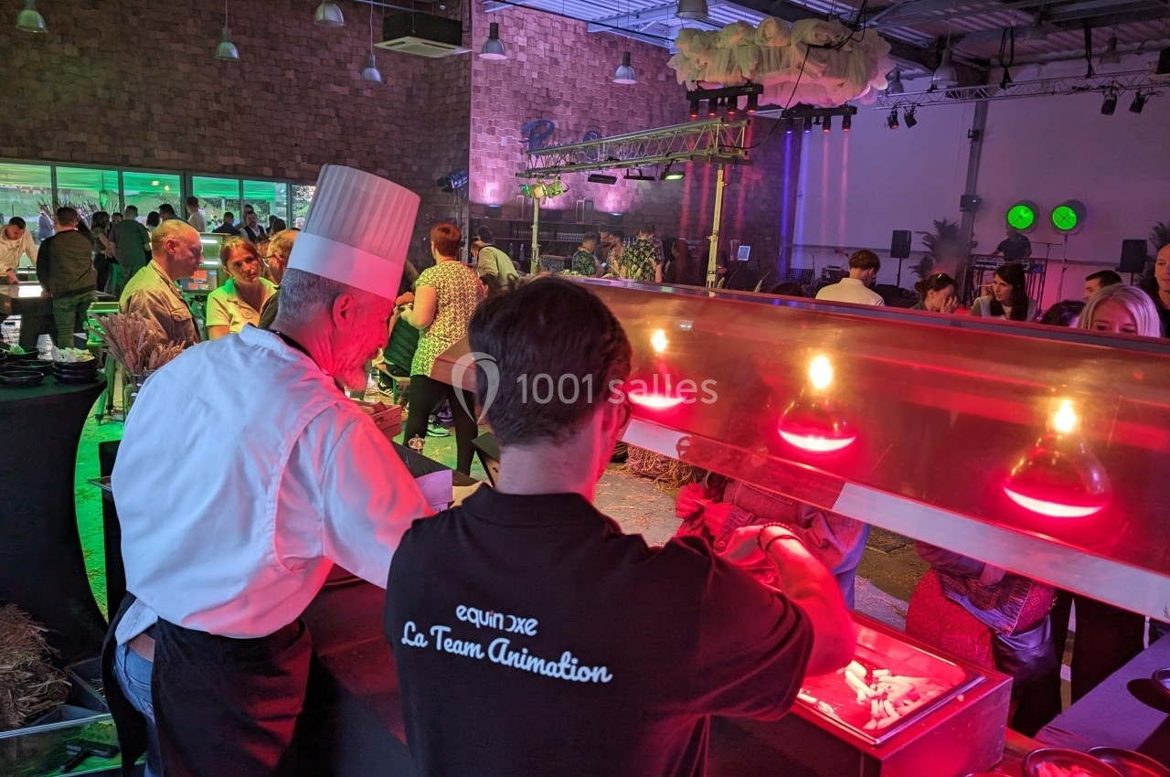 Un chef et un serveur préparent des plats sous des lampes chauffantes lors d'un événement avec des invités en arrière-plan.