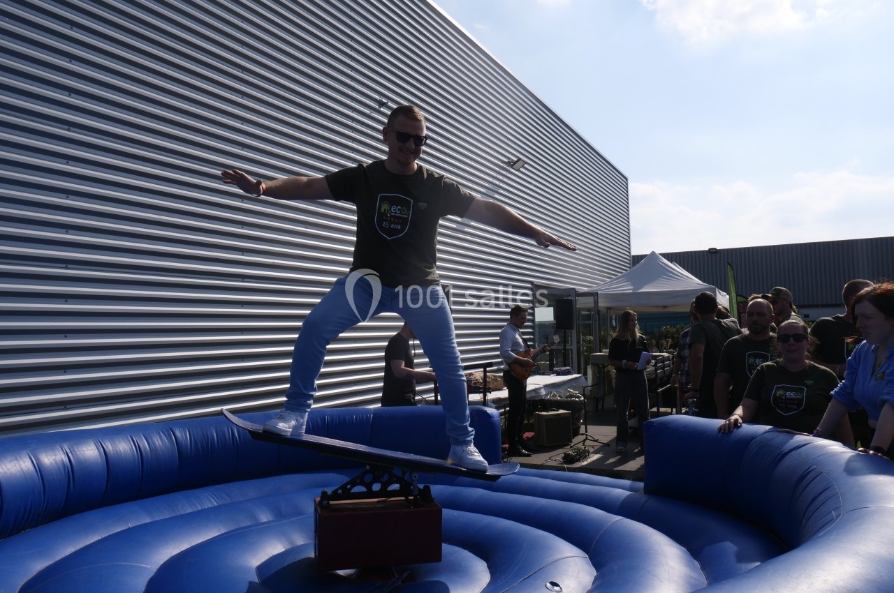 Un homme debout sur un simulateur de surf gonflable lors d'un événement en extérieur, entouré de spectateurs.