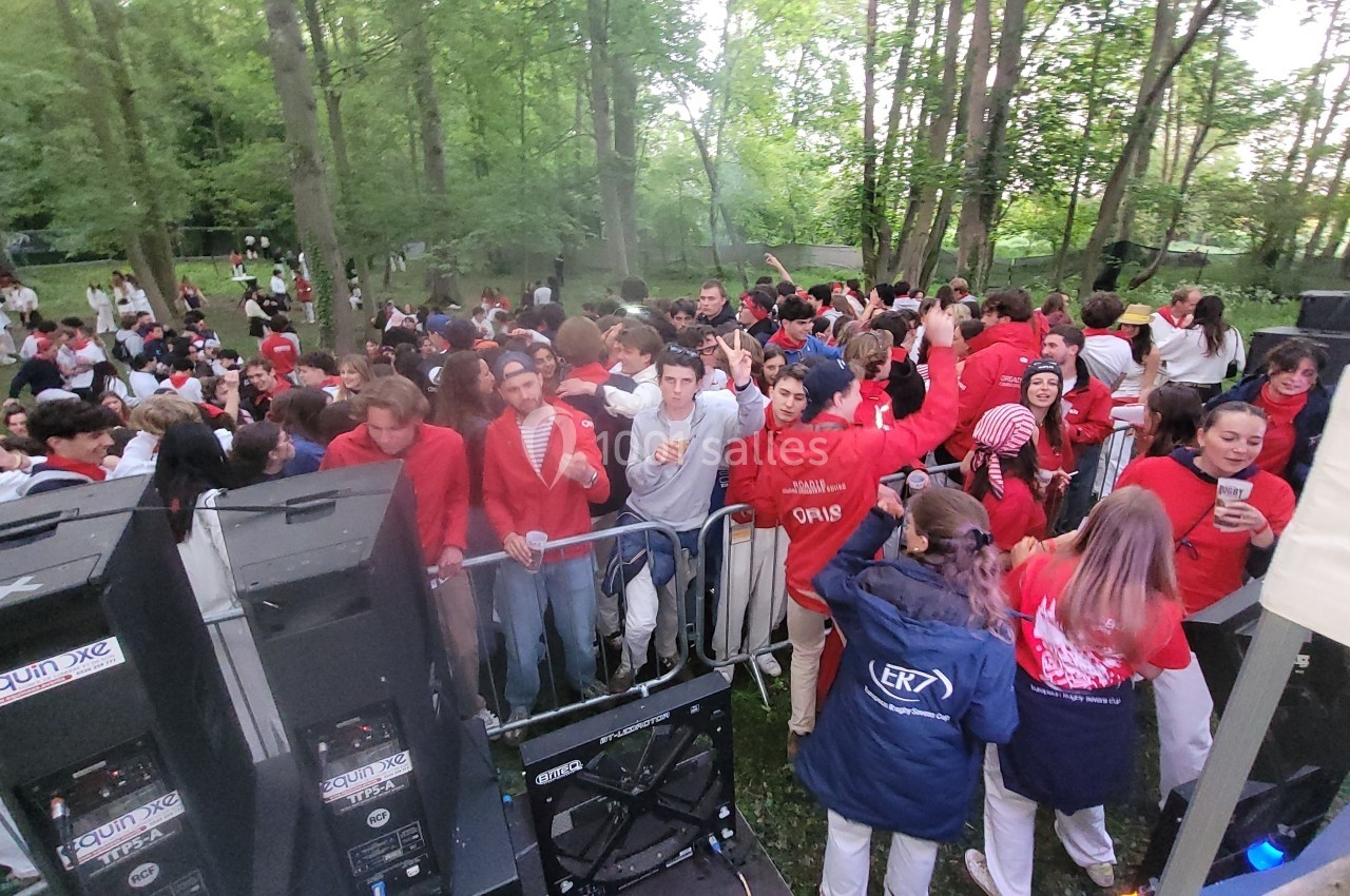 Groupe de personnes en tenue rouge et blanche dans une fête en plein air, entourées d'arbres et de matériel audio.