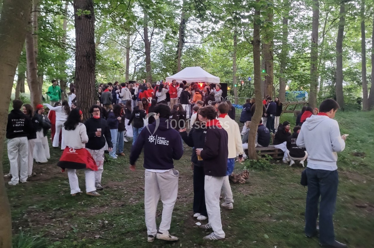 Groupe de personnes rassemblées dans un bois autour d'un stand sous une tente blanche, en tenue décontractée.