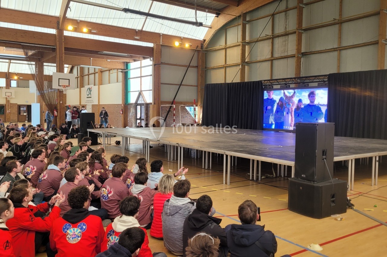 Un groupe de personnes assises dans un gymnase regarde un écran géant diffusant une vidéo sur une scène.