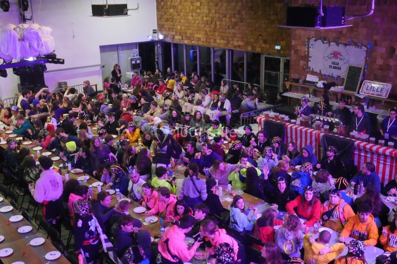 Salle bondée avec des tables alignées, des convives en tenue colorée et un stand de service en arrière-plan.