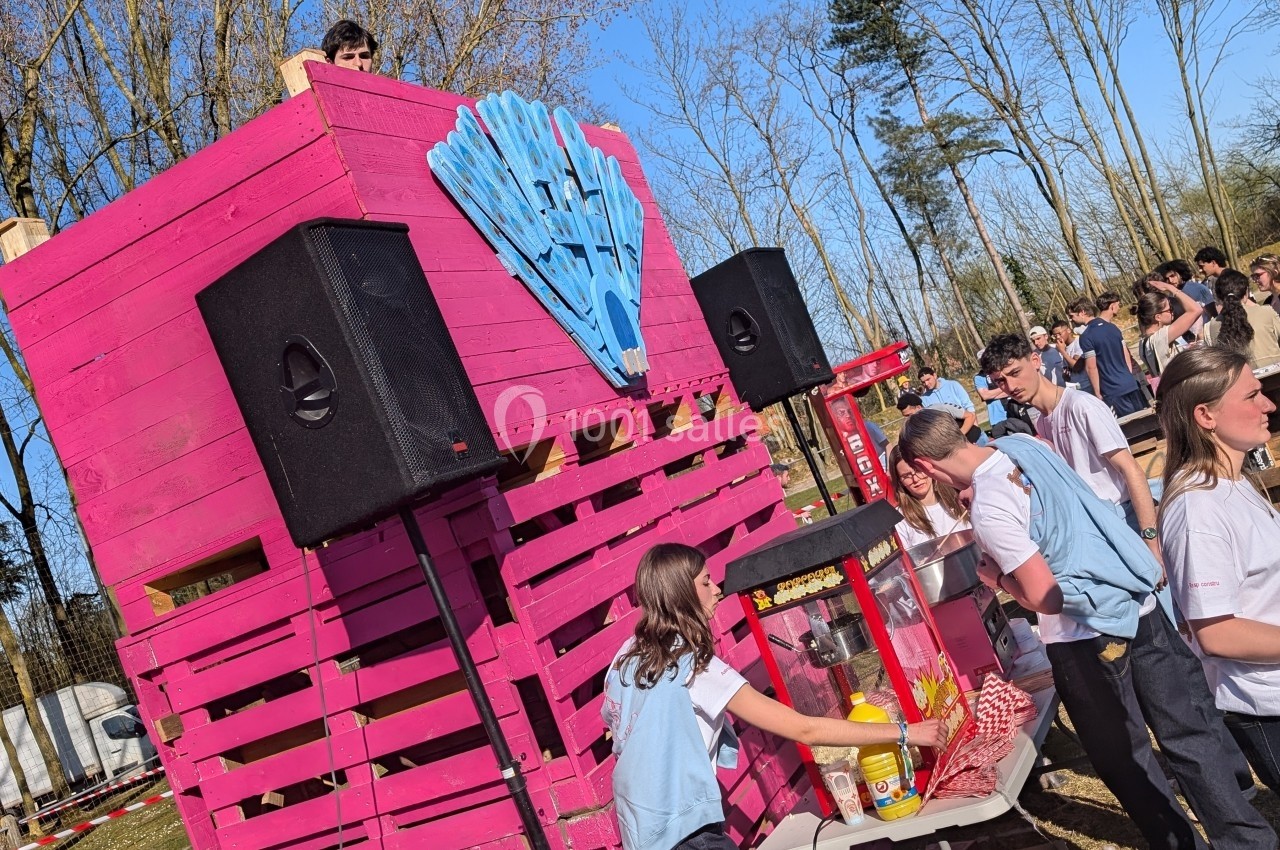 Stand en bois rose avec des haut-parleurs, une machine à popcorn et des personnes en tenue claire servant des snacks.