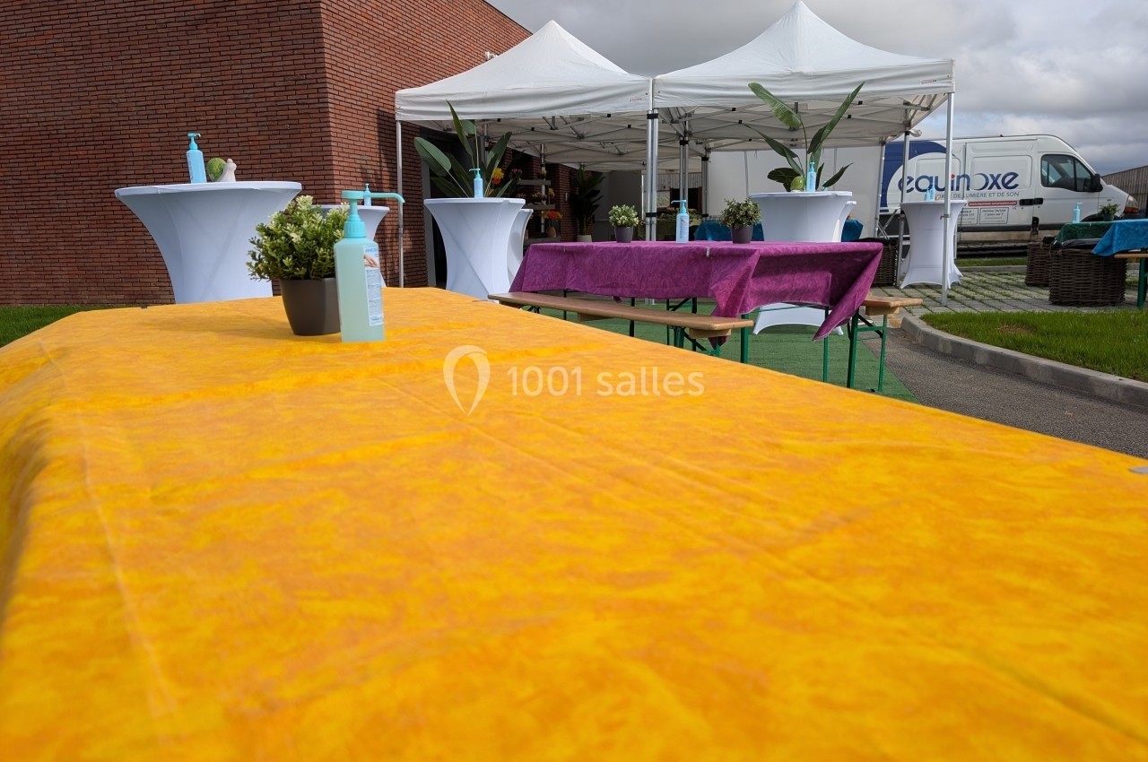 Tables recouvertes de nappes colorées avec plantes et gel hydroalcoolique, sous des tentes blanches en extérieur.