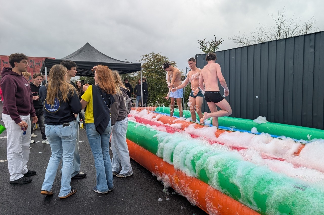 Des jeunes hommes en maillot de bain participent à une activité sur une structure gonflable recouverte de mousse, entourés…