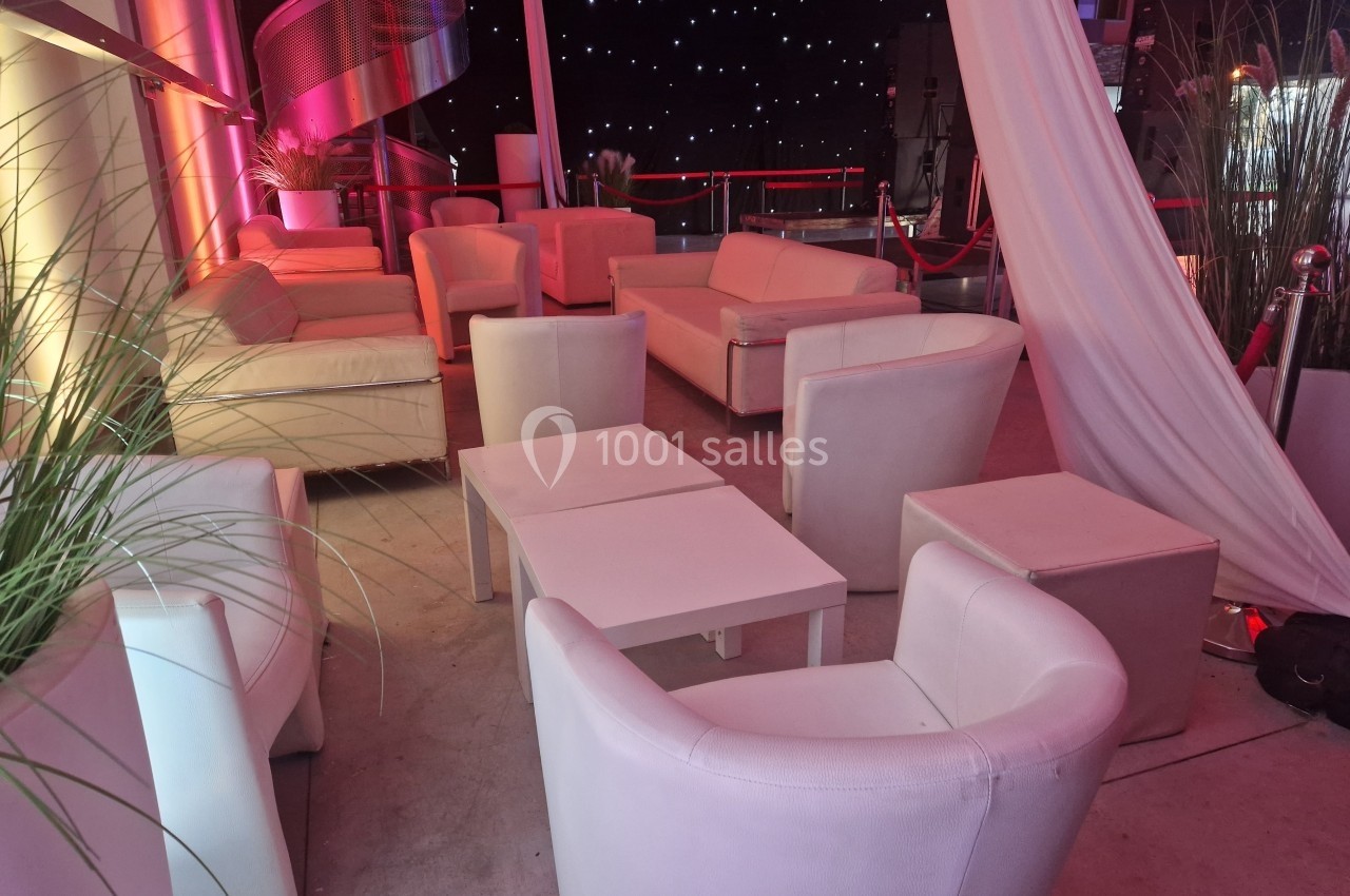 Salon avec fauteuils et canapés blancs disposés autour de petites tables, éclairage tamisé et rideaux blancs.