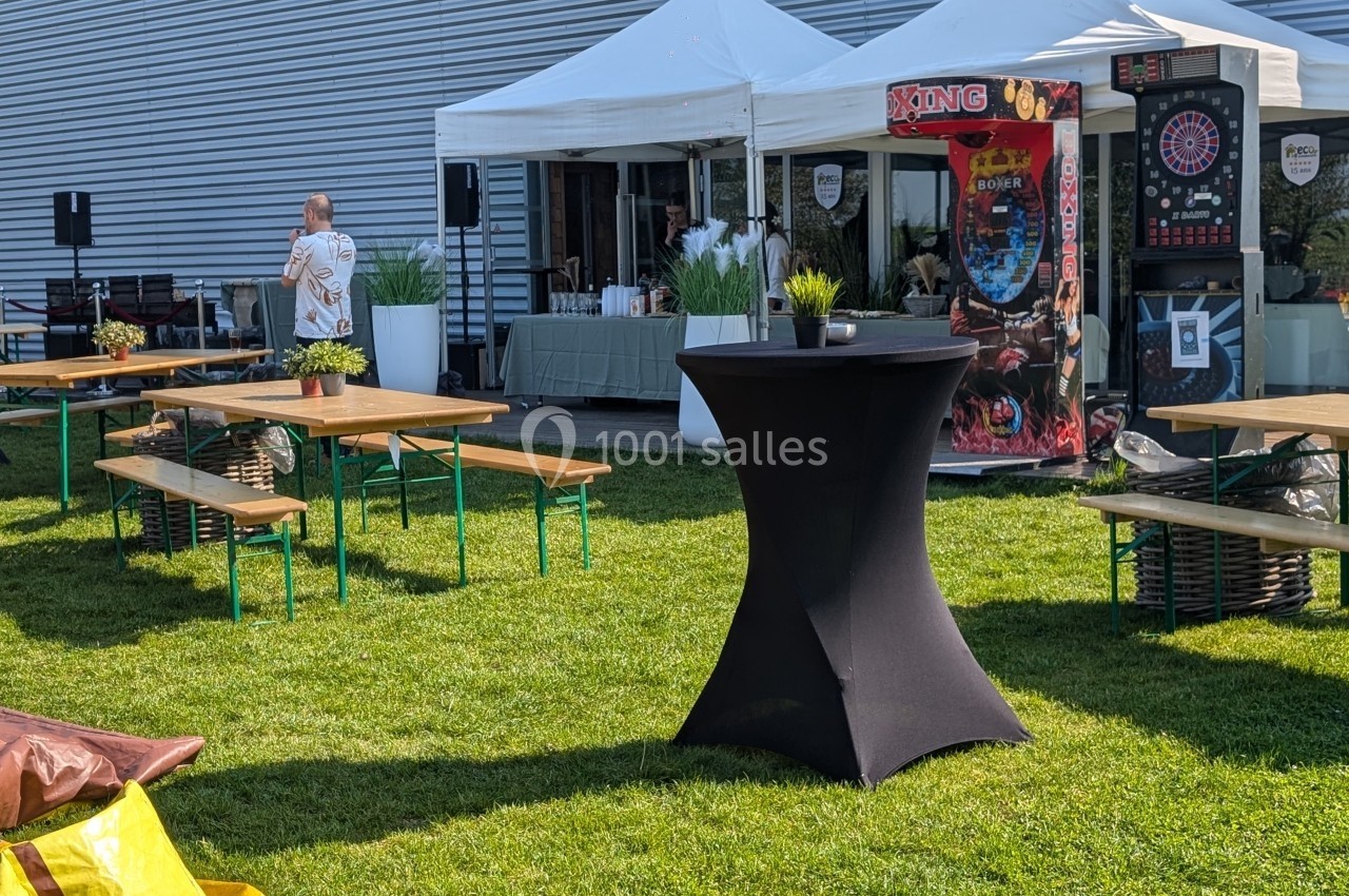 Tables et chaises en bois disposées sur une pelouse, avec des stands et jeux sous des tentes blanches en arrière-plan.