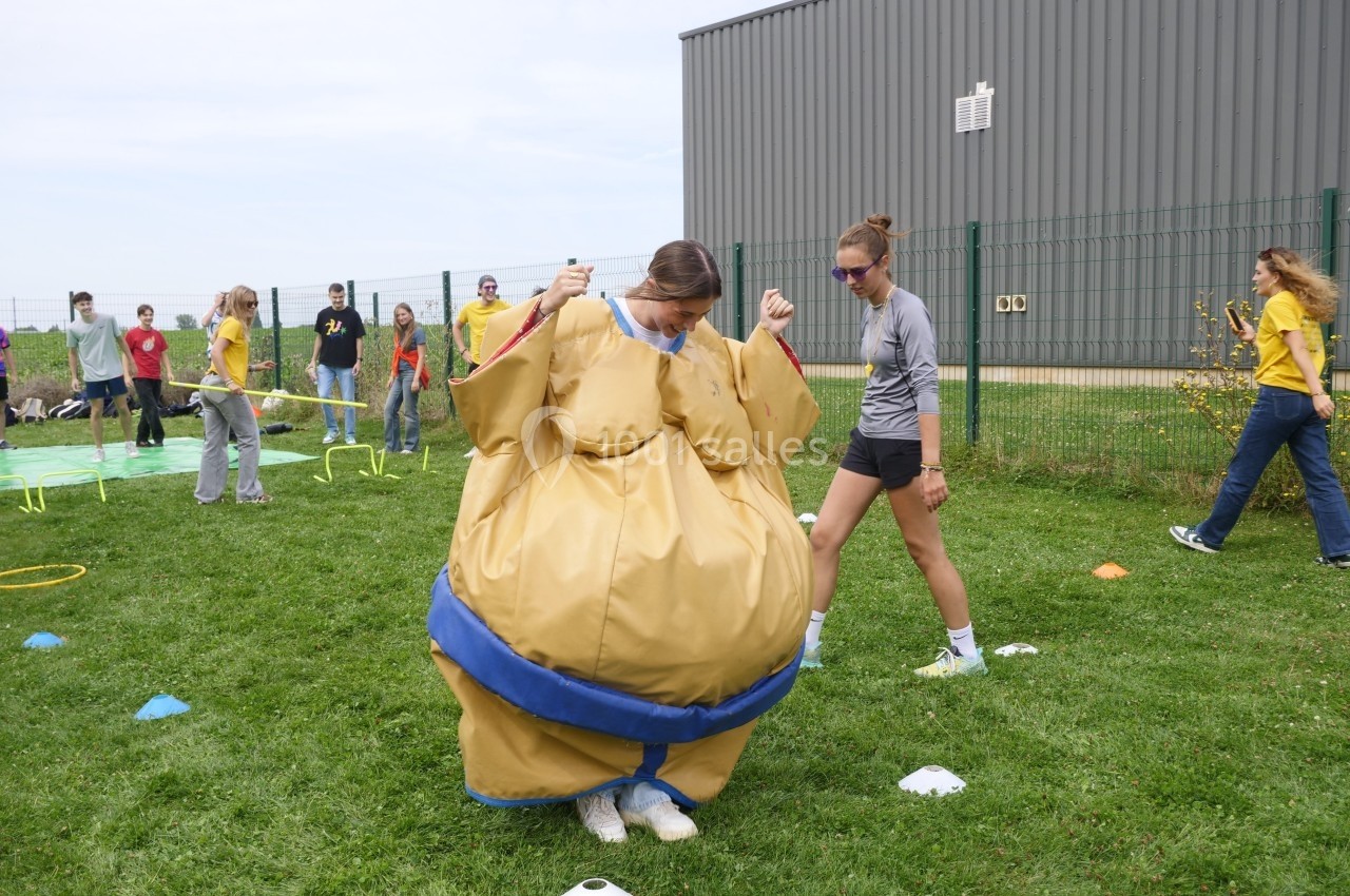 Une personne en costume de sumo avance sur une pelouse lors d'une activité en plein air, entourée de spectateurs.