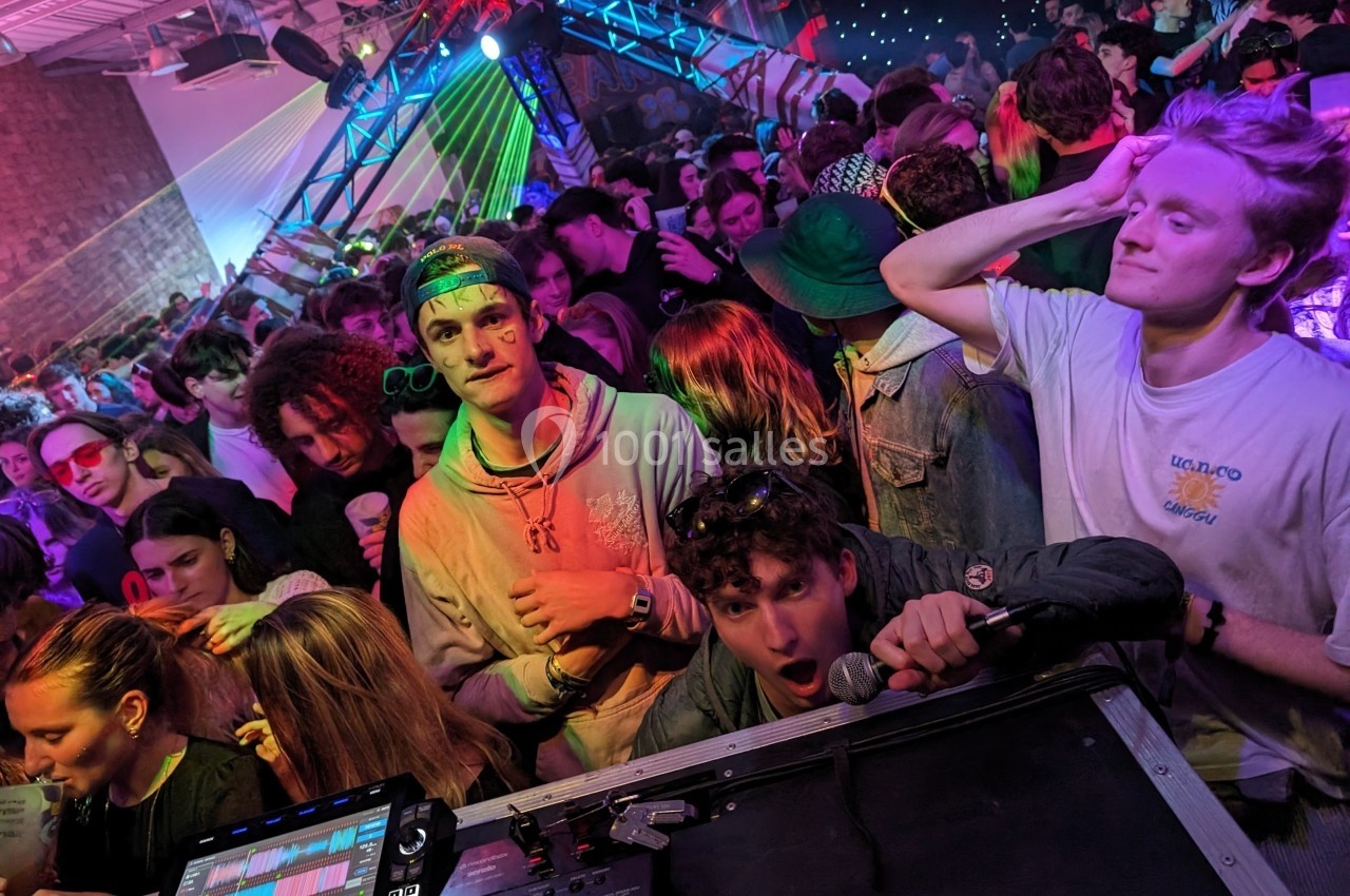 Foule animée dans une soirée festive avec un DJ en arrière-plan et des jeux de lumières colorées.