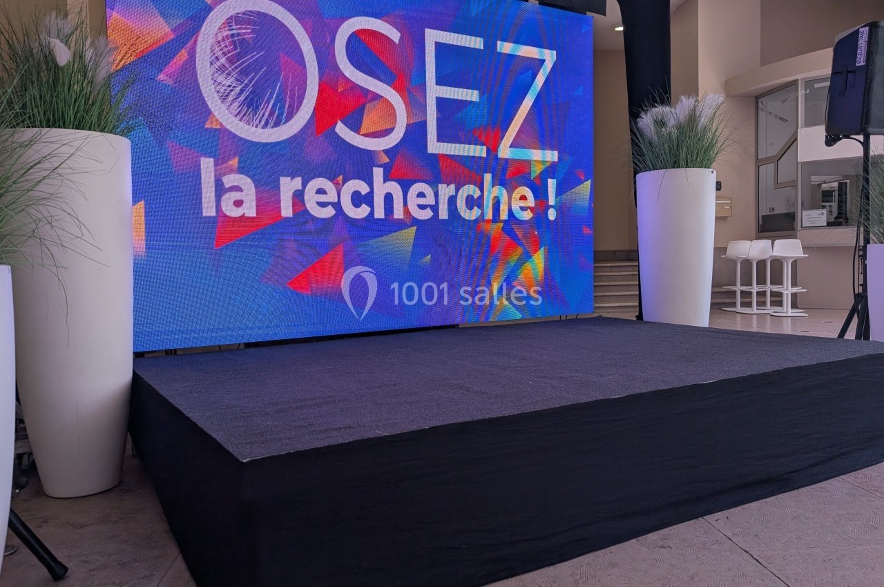Écran coloré sur une scène avec le message ’Osez la recherche !’ entouré de plantes décoratives.