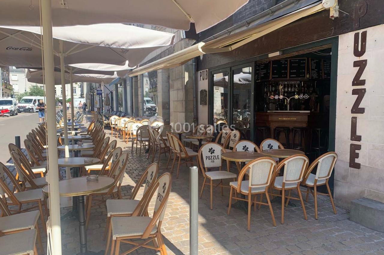 Terrasse d’un café avec des chaises en rotin et des tables sous des parasols, située le long d’une rue pavée.