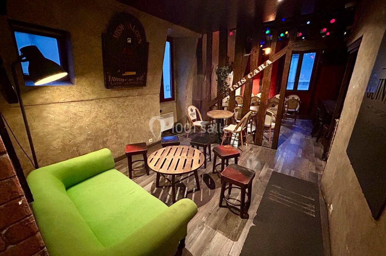 Salle chaleureuse avec canapé vert, tables en bois, chaises et éclairage tamisé, ambiance conviviale.
