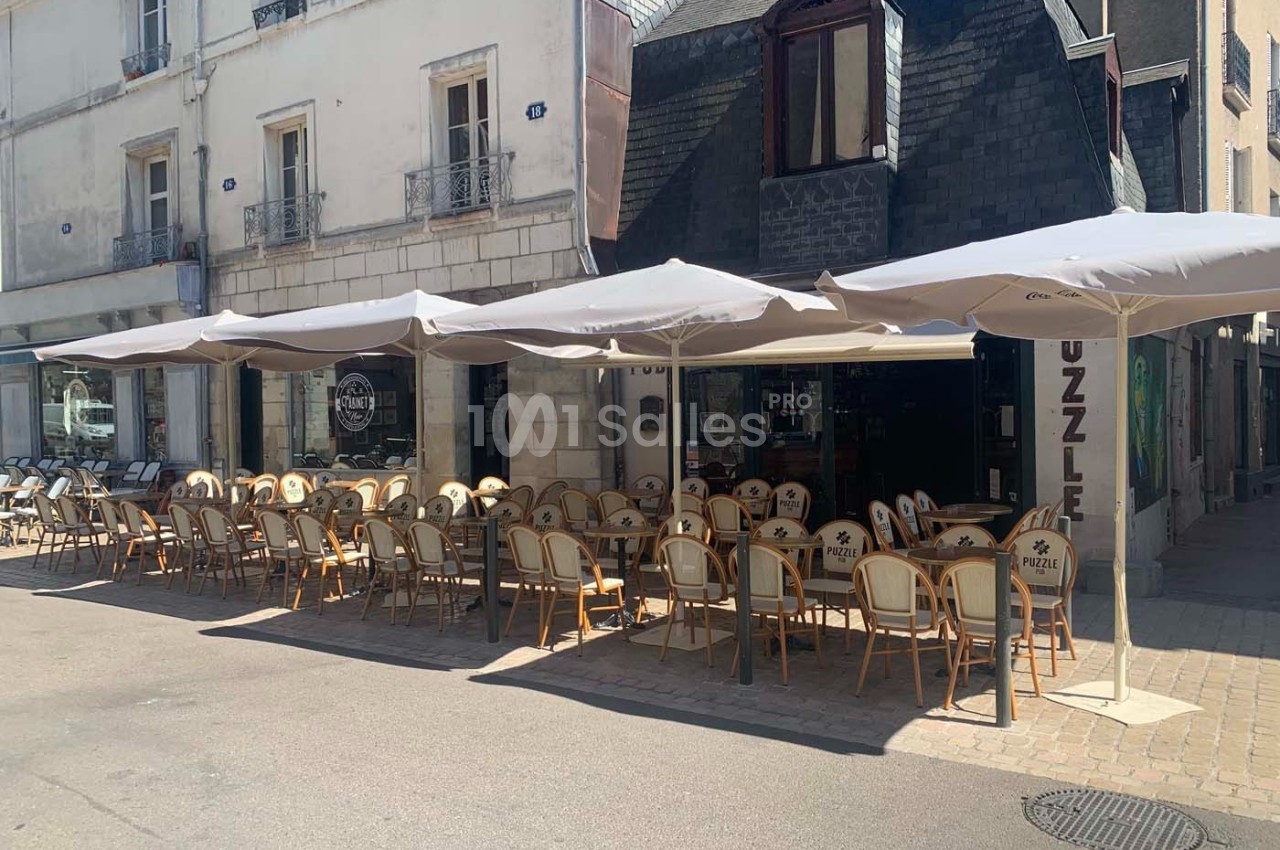 Terrasse d'un café-restaurant avec chaises en rotin et parasols, située dans une rue piétonne ensoleillée.