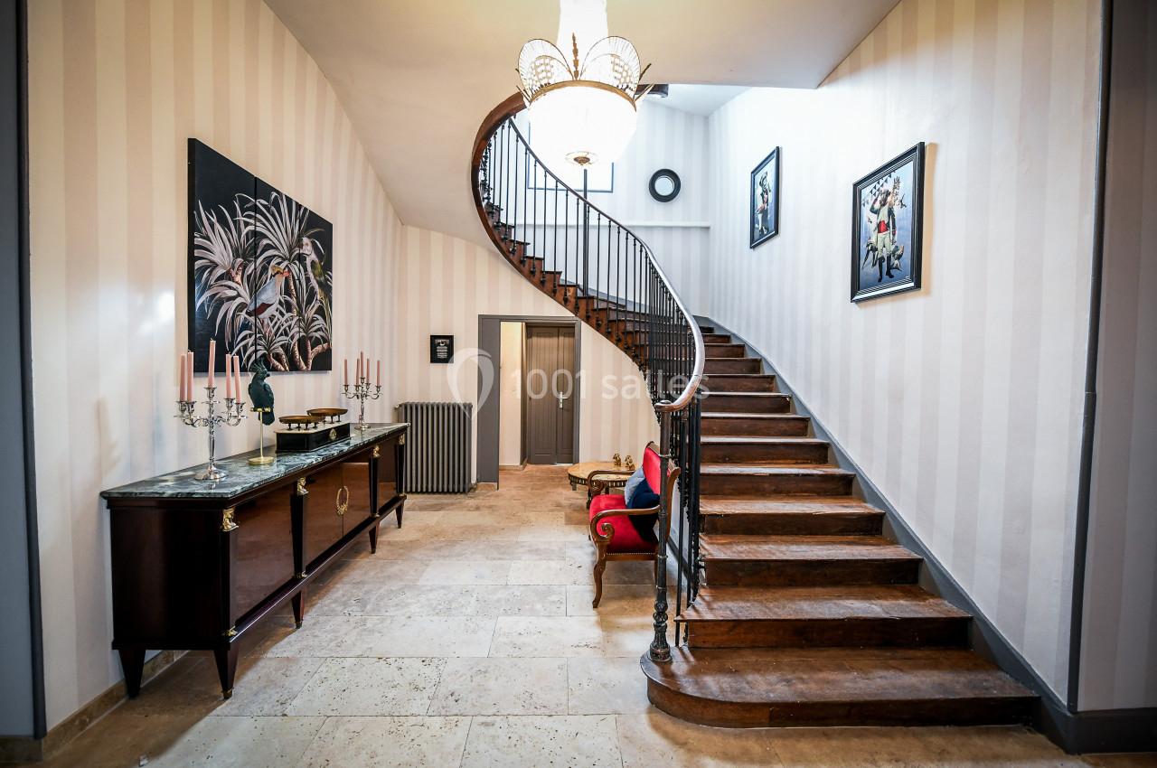 Escalier en bois courbé dans un hall lumineux avec un meuble ancien, des tableaux et un lustre suspendu.