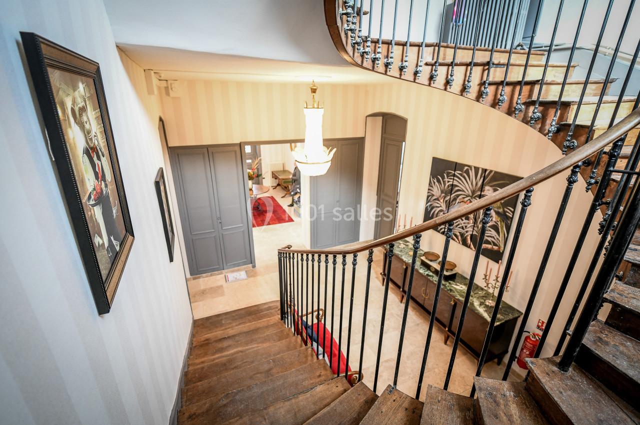 Escalier en bois avec rampe en fer forgé, menant à un intérieur décoré de tableaux et meubles classiques.