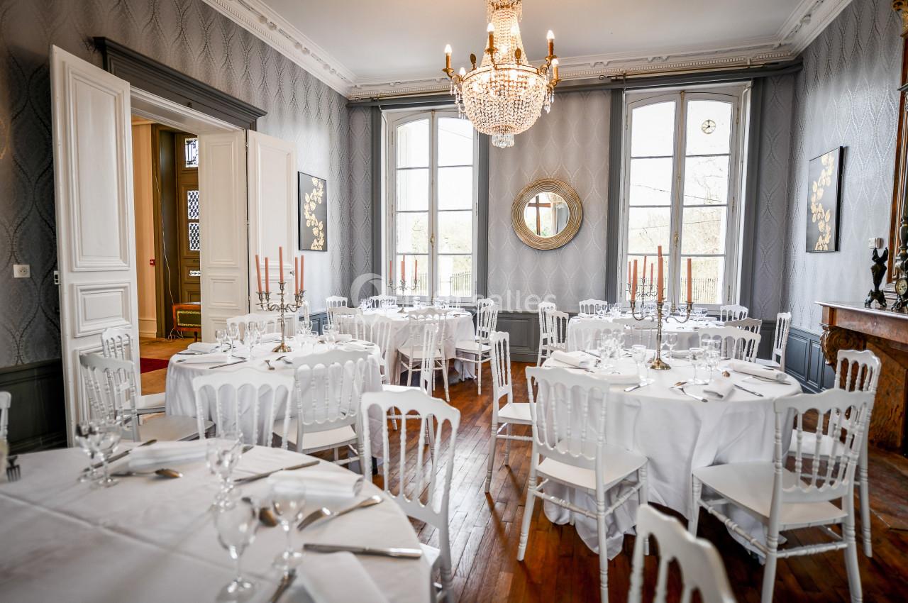 Salle élégante avec tables rondes dressées, chaises blanches, chandeliers et lustre, éclairée par de grandes fenêtres.