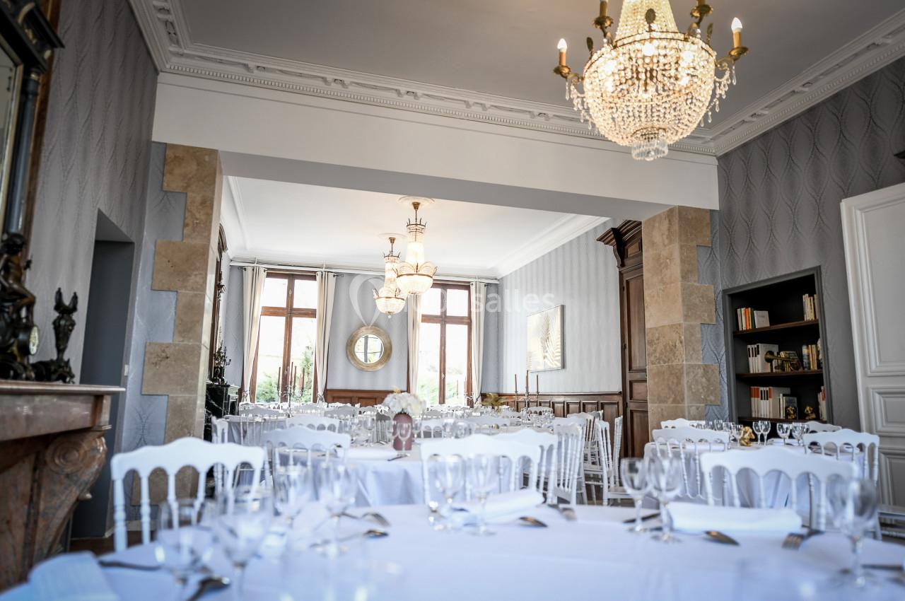 Salle élégante avec tables dressées, chaises blanches, lustres en cristal et lumière naturelle entrant par les fenêtres.