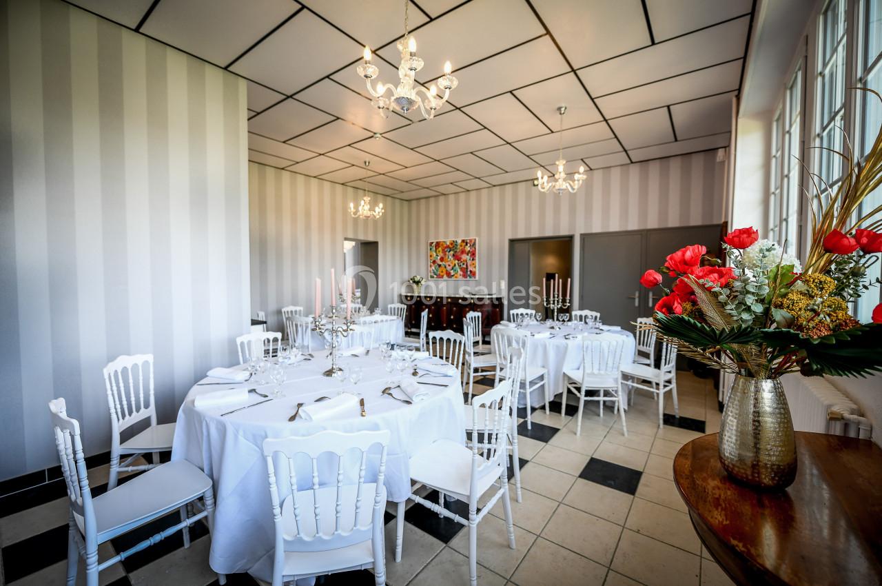 Salle lumineuse avec tables rondes dressées, chaises blanches, lustres et décoration florale dans un cadre élégant.