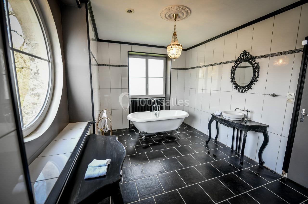 Salle de bain élégante avec baignoire sur pieds, carrelage noir, miroir orné et grande fenêtre ronde.