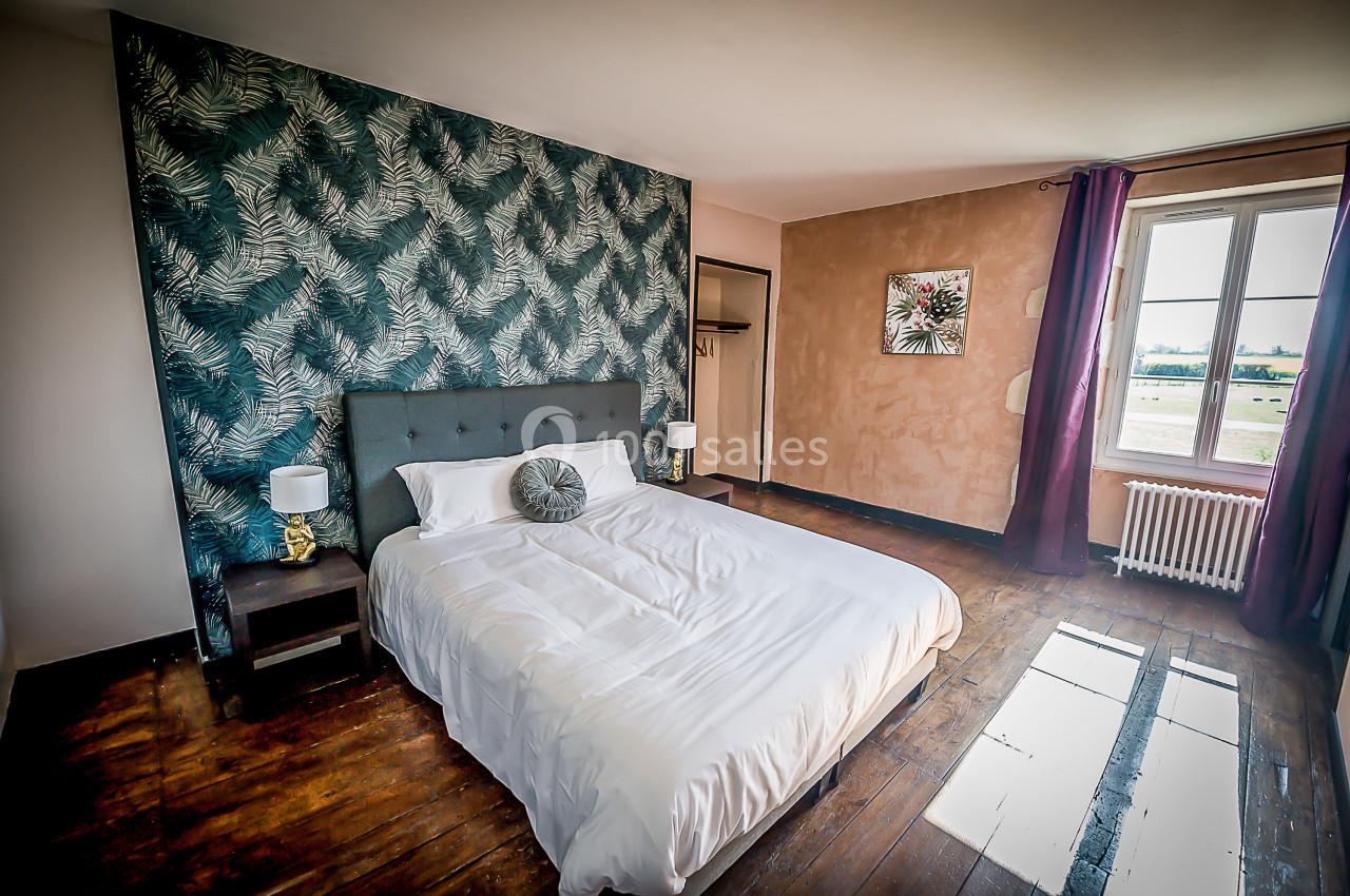 Chambre avec lit double, mur décoré de motifs végétaux, parquet en bois et fenêtre laissant entrer la lumière naturelle.