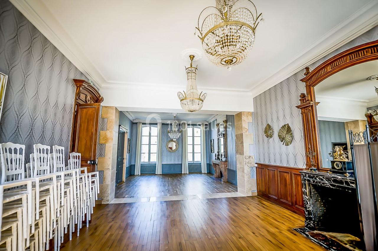 Salle élégante avec parquet, murs ornés de boiseries et miroirs, lustres en cristal et chaises empilées sur le côté.