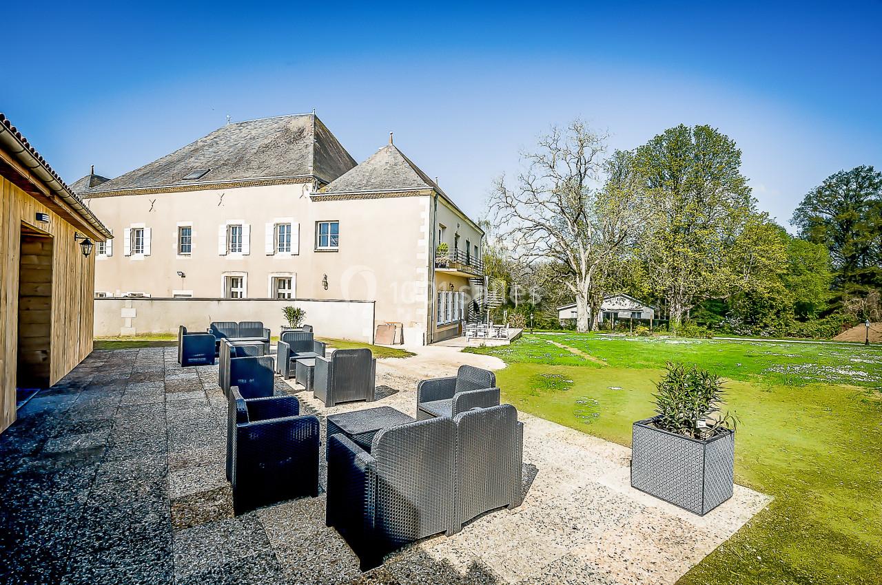 Terrasse aménagée avec fauteuils en rotin, donnant sur un jardin verdoyant et un bâtiment ancien sous un ciel bleu.