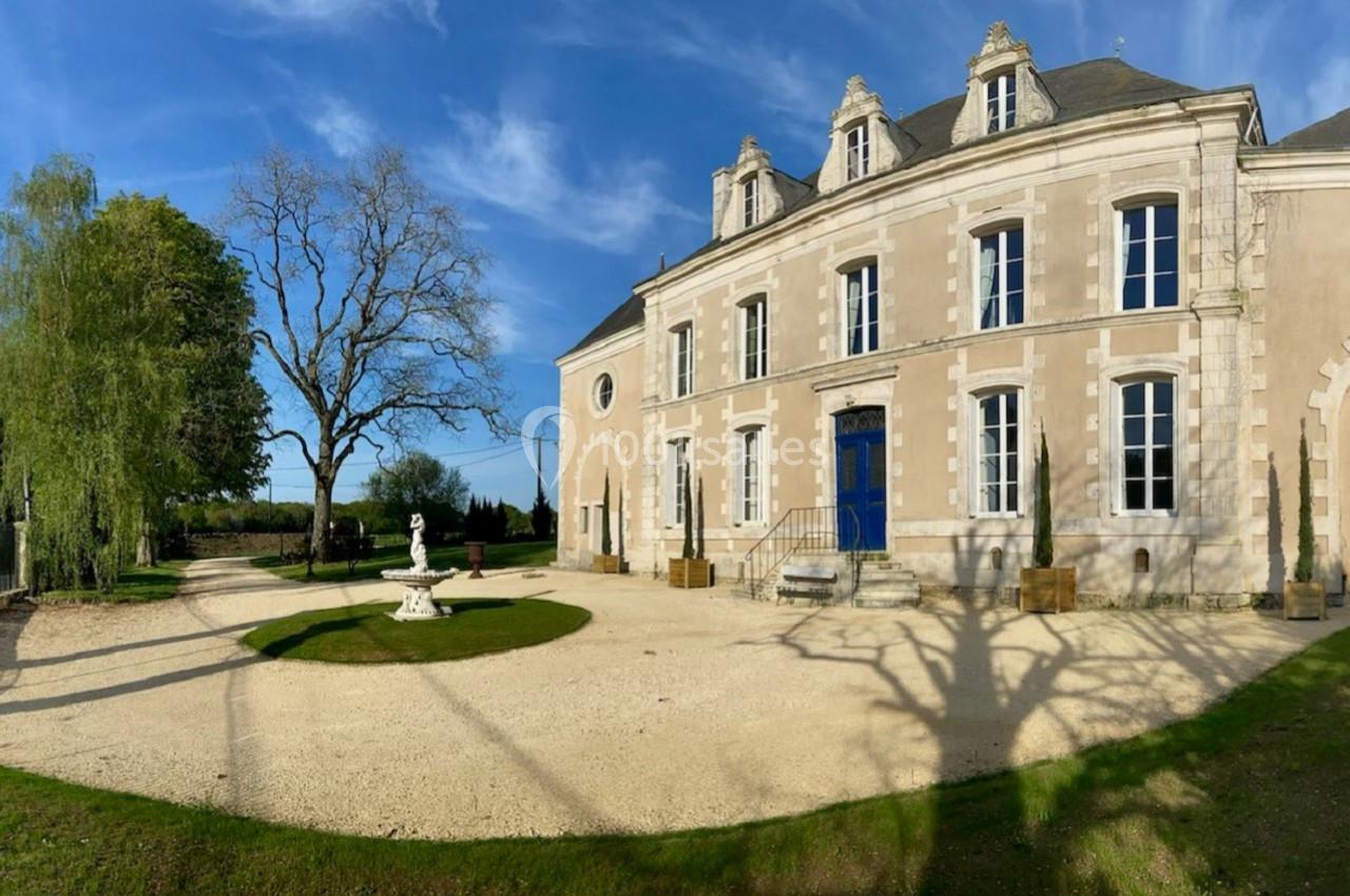 Façade d'un manoir en pierre avec une porte bleue, entouré d'un jardin arboré et d'une allée gravillonnée.