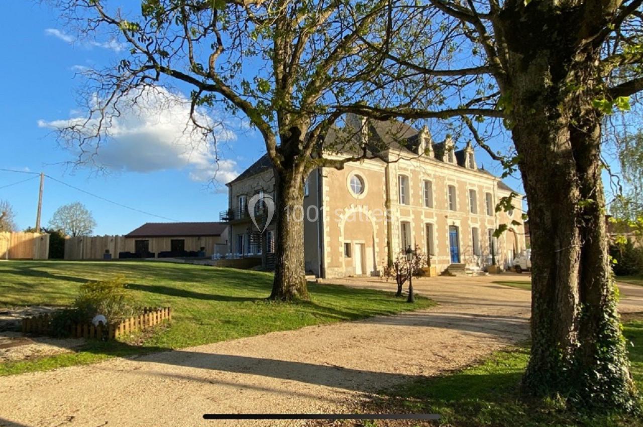 Grande maison en pierre avec volets bleus, entourée d'arbres et d'une pelouse, sous un ciel dégagé.