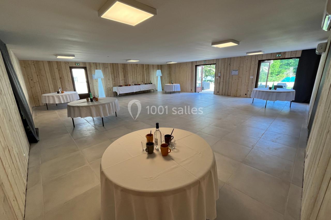 Salle lumineuse avec murs en bois, tables rondes et rectangulaires dressées, vue sur l'extérieur par des baies vitrées.