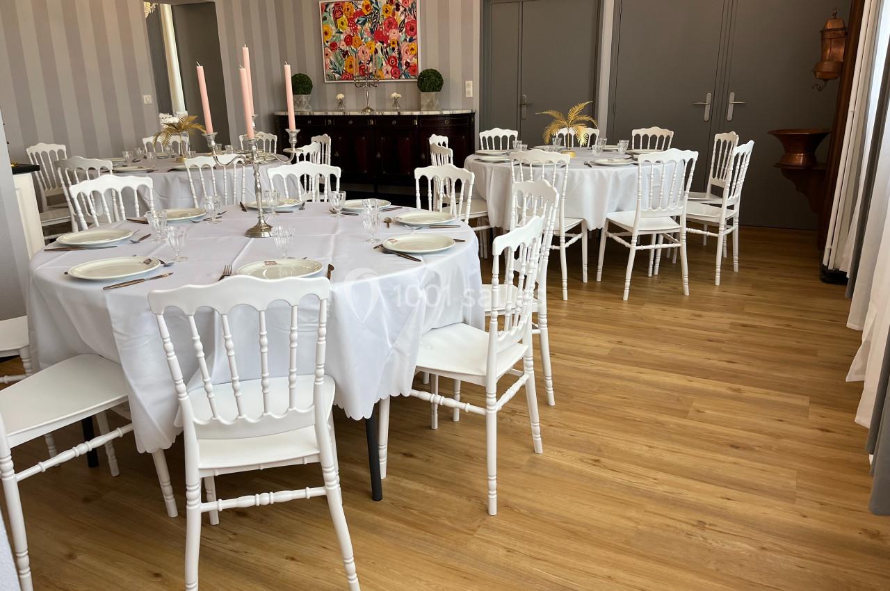 Salle décorée avec des tables rondes dressées, nappes blanches, chaises blanches et bougeoirs, sur un sol en bois clair.