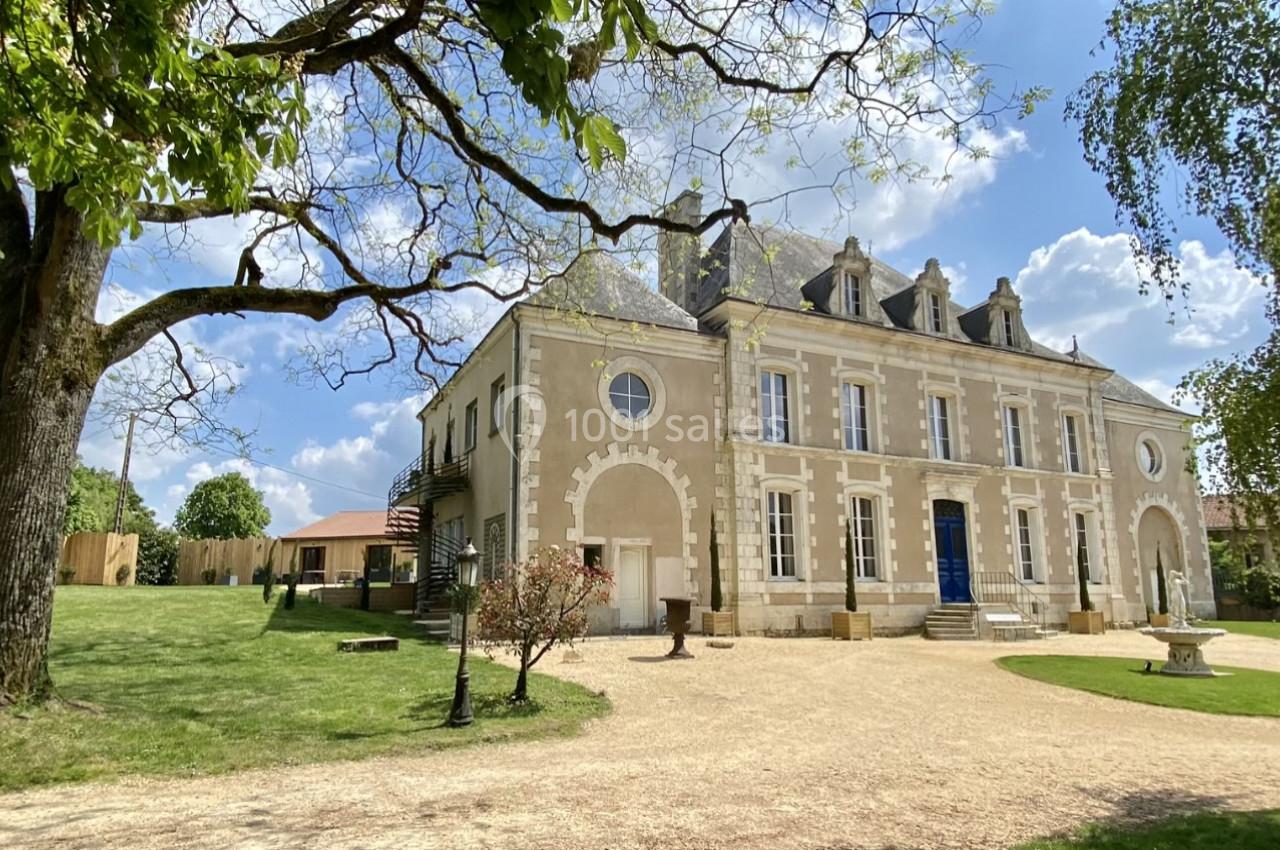 Façade d'une grande maison en pierre entourée d'un jardin arboré sous un ciel bleu avec quelques nuages.