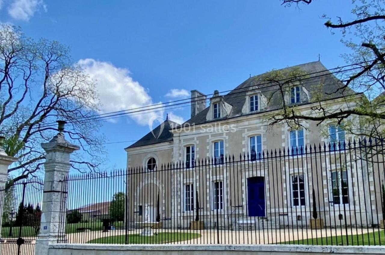 Façade d'un grand bâtiment en pierre, entouré d'une grille en fer forgé et d'arbres, sous un ciel bleu.