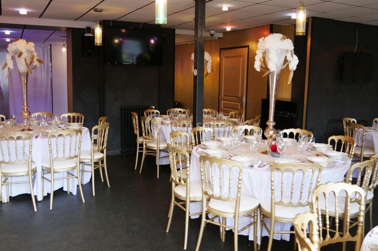Salle de réception décorée avec des tables rondes, nappes blanches, chaises dorées et centres de table fleuris.
